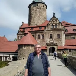 Mężczyzna w okularach i niebieskiej kamizelce na tle kamiennego zamku z wieżą i czerwonym dachem w słoneczny dzień.