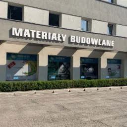 Fasada budynku składu budowlanego Sanier z dużym napisem 'Materiały Budowlane' oraz reklamami producentów Bostik, Kabe i Schomburg w oknach wystawowych.
