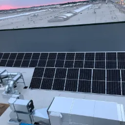 Instalacja fotowoltaiczna na dachu budynku przemysłowego z widocznymi panelami słonecznymi, instalacją wentylacyjną i fragmentem różowego nieba o zmierzchu w tle.