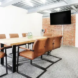 Nowoczesna sala konferencyjna z drewnianym stołem, brązowymi skórzanymi krzesłami, ceglaną ścianą i wiszącym telewizorem.