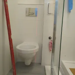 Montaż podwieszanej toalety z użyciem poziomicy laserowej, widoczna kabina prysznicowa i białe płytki na ścianach.