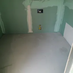 Surowe wnętrze łazienki z zielonymi płytami kartonowo-gipsowymi, przygotowane do dalszych prac wykończeniowych, widoczne instalacje wodno-kanalizacyjne i otw&oacute;r rewizyjny.