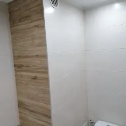 Wykończona łazienka z białymi płytkami i panelami imitującymi drewno, widoczny fragment toalety i kratka wentylacyjna.