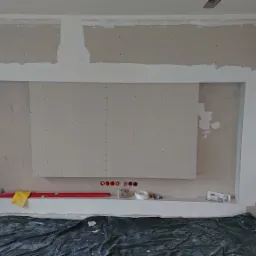 Wnętrze w trakcie prac w Białymstoku: zamontowane płyty na ścianie, widoczne ślady po szpachlowaniu i przygotowaniu pod malowanie, podłoga zabezpieczona folią, poziomica i narzędzia na dolnej półce.