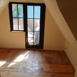 Poddasze z drewnianą podłogą i oknem balkonowym, widoczne skosy ścian i drewniana boazeria sufitowa, wpadające promienie słoneczne podkreślają strukturę drewna.