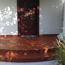 Wejście do domu z brązowymi płytkami na schodach i podłodze, białe kolumny i ciemne drewniane drzwi, na ścianie gra świateł i cieni, w rogu widoczne ozdobne trawy.