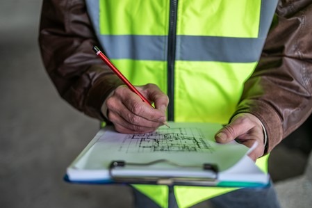 Osoba w kamizelce kreśli plan budynku na papierze z czerwoną kredką. Ręce i fragment ubrania. Plan architektoniczny na białym tle.