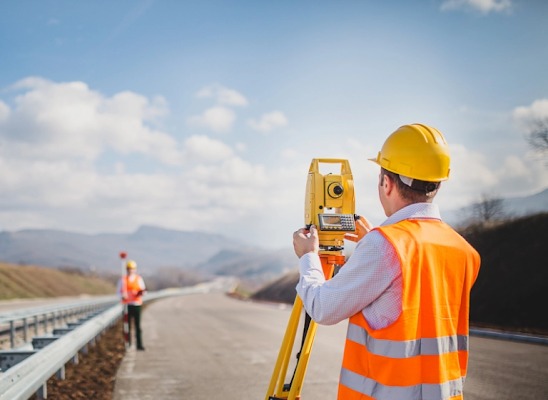 Praca geodezyjna na drodze: geodeta w kasku i kamizelce mierzy niwelatorem, w tle drugi geodeta z łatą niwelacyjną. Precyzyjne pomiary w budownictwie drogowym.