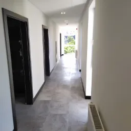 Długi, jasny korytarz z szarą podłogą wyłożoną panelami imitującymi kamień, czarne obramowania drzwi, białe ściany i widok na zieleń na końcu korytarza.