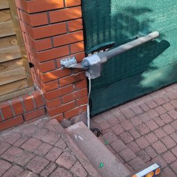 ELEKTROSMART - Zamontowany siłownik automatyki bramy na ceglanym słupku, widoczny poziomica i narzędzia na kostce brukowej. Zielona siatka w tle.