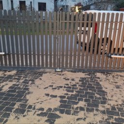 ELEKTROSMART - Szara, metalowa brama posesyjna z automatyką, widoczna z zewnątrz na tle zaparkowanego białego busa. Kostka brukowa przed bramą pokryta piaskiem.