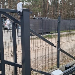ELEKTROSMART - Szara, metalowa brama przesuwna z automatyką, widoczny napęd i fotokomórka. W tle drzewa i fragment ogrodzenia panelowego. Brama na posesji.