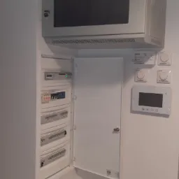 Otwarta szafka rozdzielcza z widocznymi bezpiecznikami i elementami sterującymi, umieszczona pod metalową szafką z szybą i kluczykiem, obok panelu sterowania ogrzewaniem i wideodomofonu.