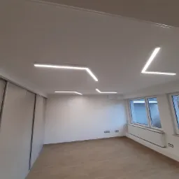 Nowoczesne oświetlenie sufitowe LED w pustym, jasnym pokoju z przesuwnymi drzwiami szafy i trzema oknami, ukazujące geometryczne kształty lamp.