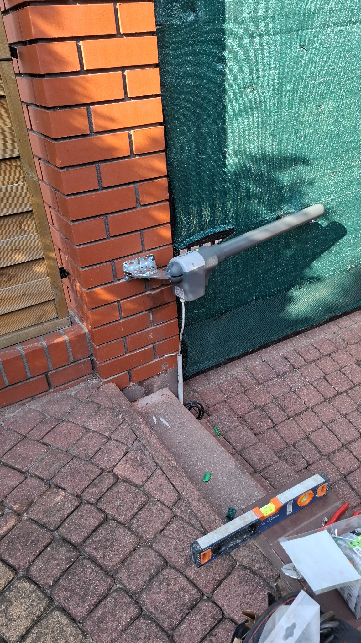 Zamontowany siłownik automatyki bramy na ceglanym słupku, widoczny poziomica i narzędzia na kostce brukowej. Zielona siatka w tle.