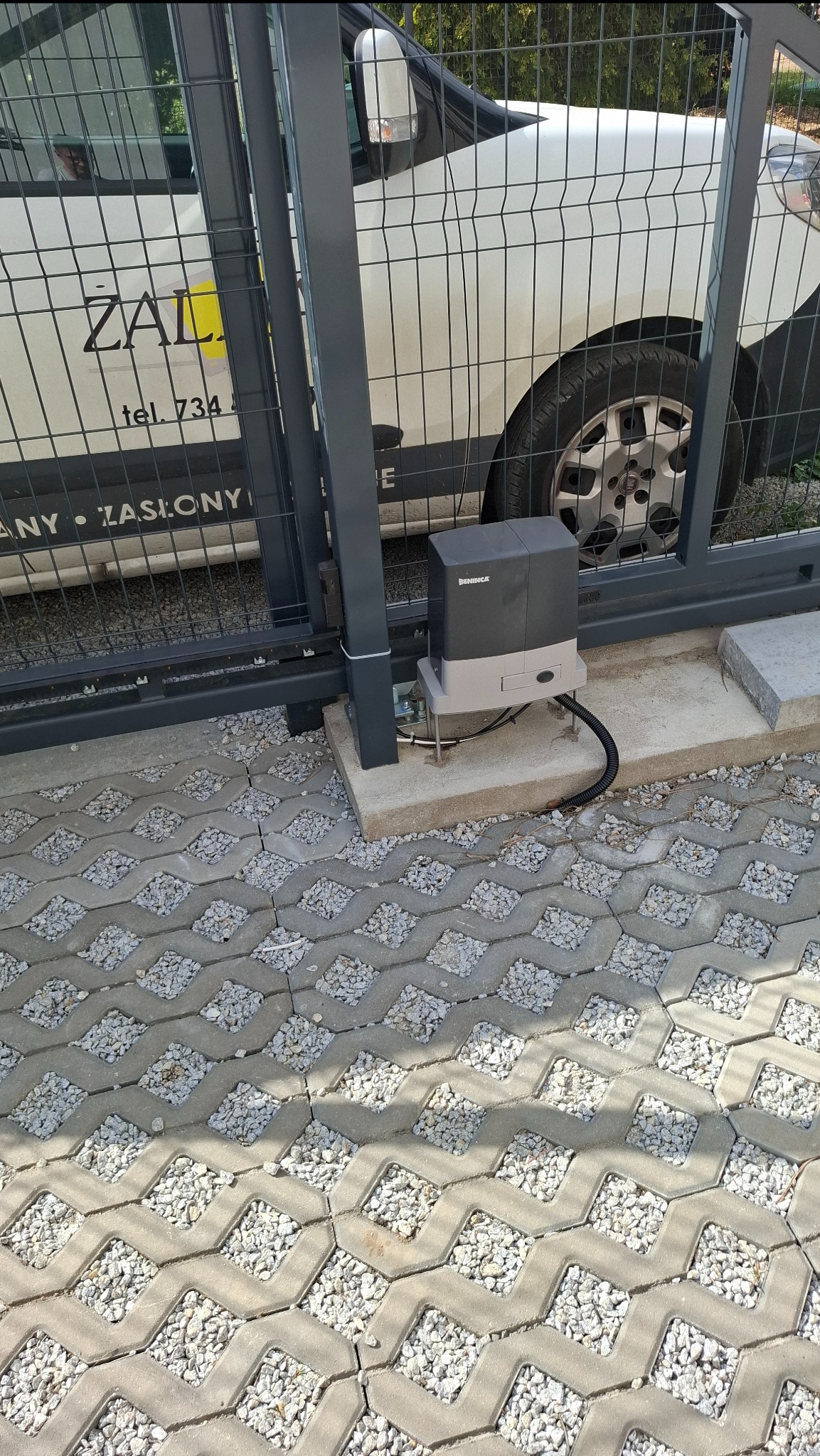 Zamontowany napęd Beninca do bramy przesuwnej na betonowej podstawie, widoczny na tle ażurowej kostki brukowej i częściowo samochodu firmy.