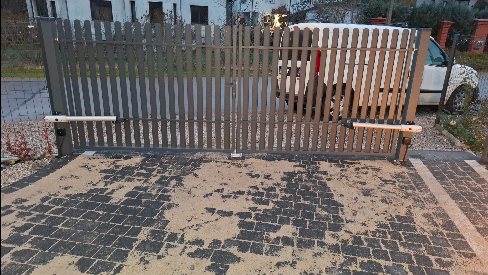 Szara, metalowa brama posesyjna z automatyką, widoczna z zewnątrz na tle zaparkowanego białego busa. Kostka brukowa przed bramą pokryta piaskiem.