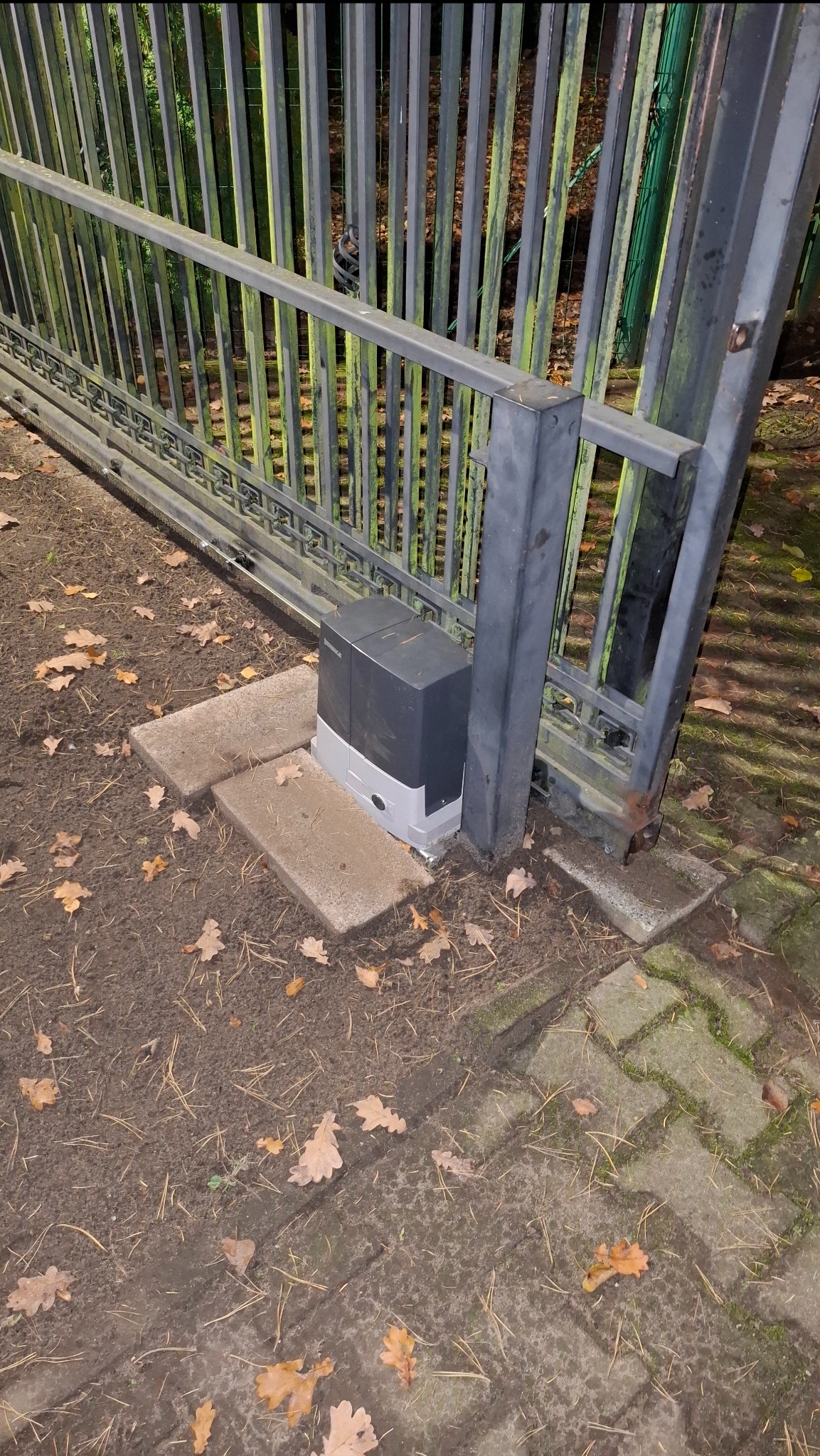 Zamontowany napęd bramy przesuwnej, widoczny na betonowej podstawie obok metalowej konstrukcji bramy. Liście na ziemi. Ujęcie z bliska, nocne oświetlenie.