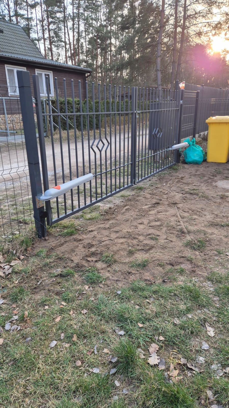Szara, metalowa brama wjazdowa z zamontowanym siłownikiem automatycznym, widoczna na tle domu i drzew. Na ziemi trawa i liście.