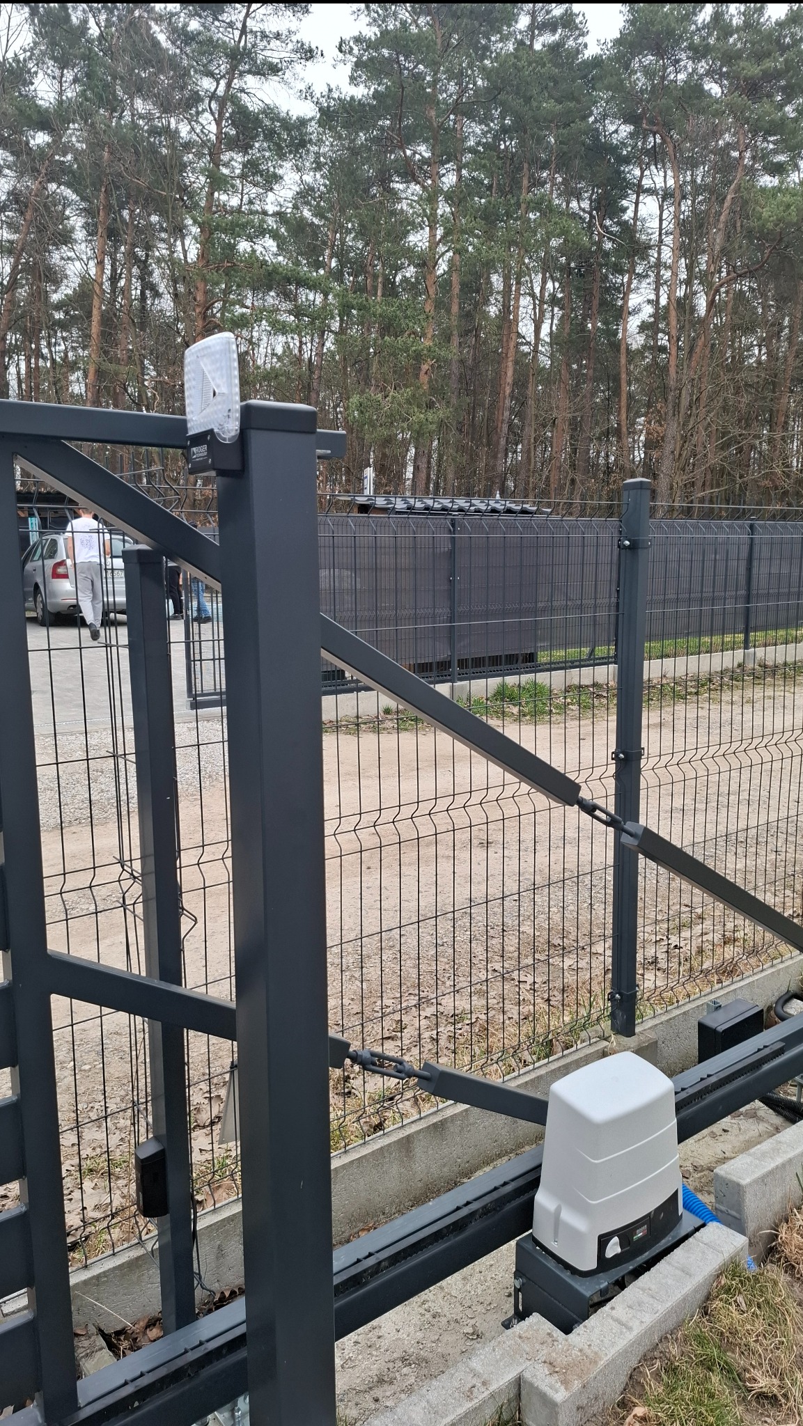Szara, metalowa brama przesuwna z automatyką, widoczny napęd i fotokomórka. W tle drzewa i fragment ogrodzenia panelowego. Brama na posesji.