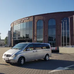 Srebrny van Mercedes-Benz klasy V zaparkowany przed Warszawskim Centrum EXPO XXI w słoneczny dzień.