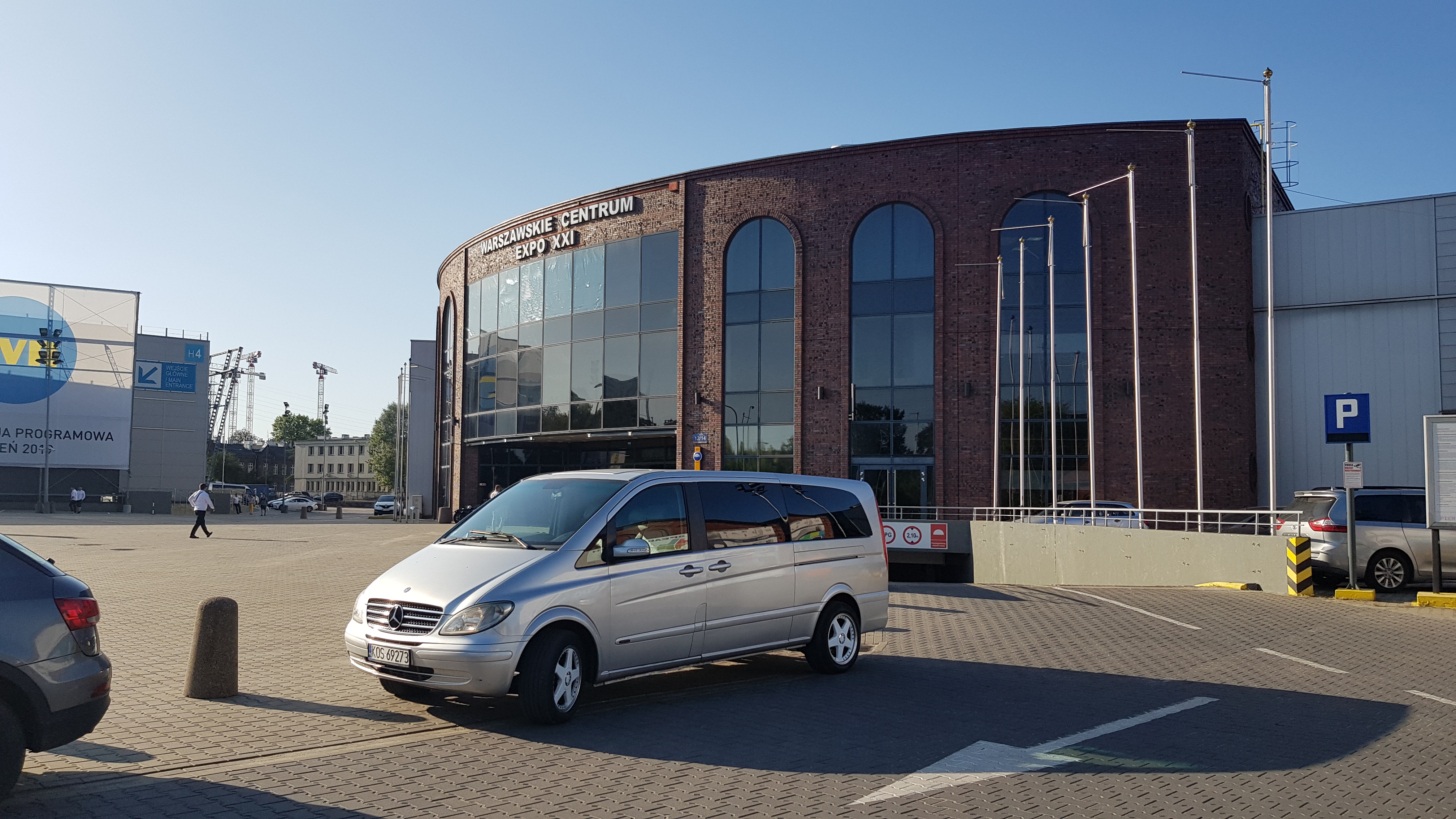 Srebrny van Mercedes-Benz klasy V zaparkowany przed Warszawskim Centrum EXPO XXI w słoneczny dzień.