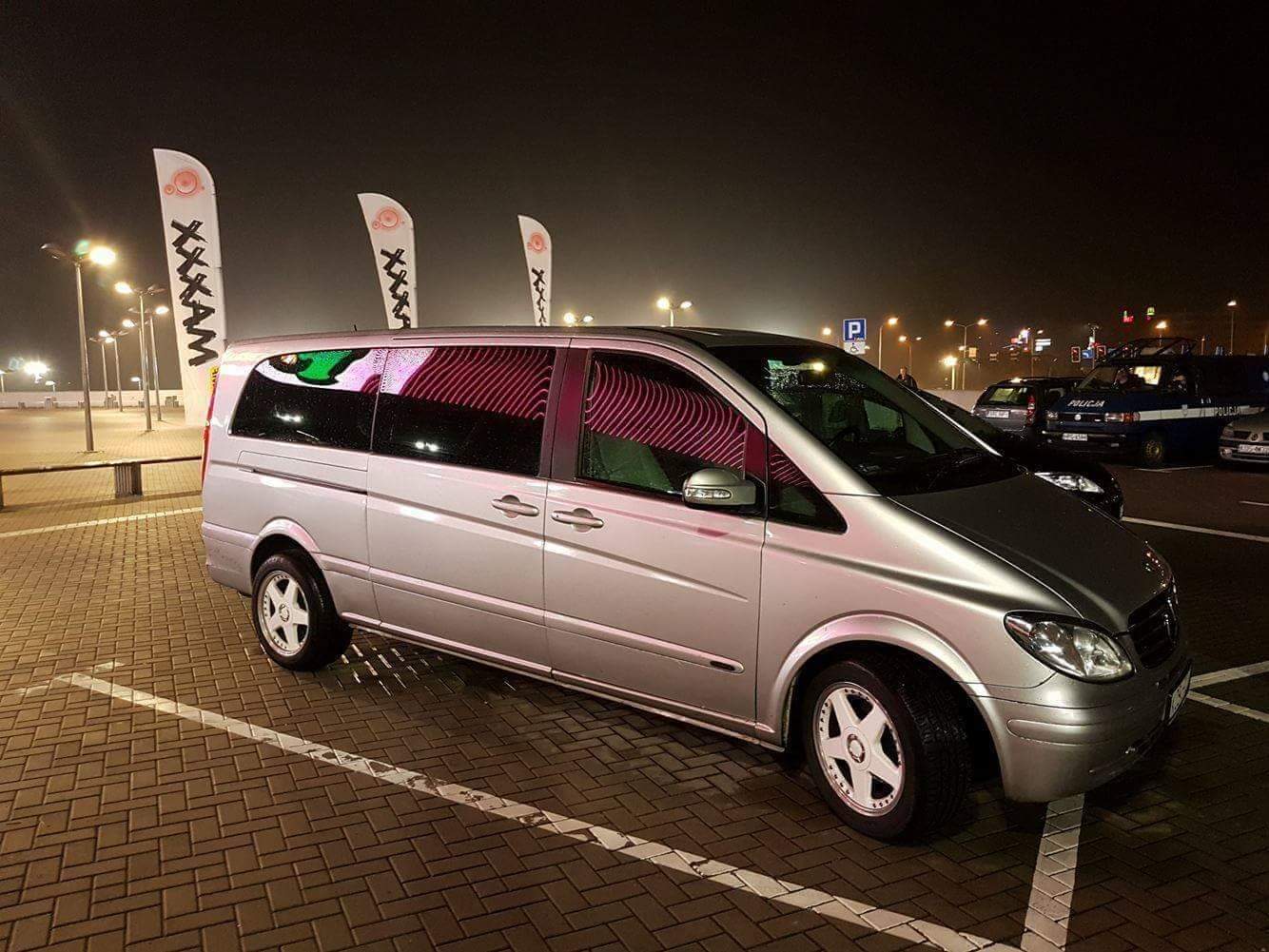 Srebrny van Mercedes Viano zaparkowany nocą na parkingu z kostki brukowej, widoczne białe flagi reklamowe i radiowóz policyjny w tle.