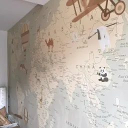 Ściana w pokoju dziecięcym pokryta tapetą z mapą świata, z ilustracjami zwierząt, samolotu i balonu na ogrzane powietrze, oraz naklejonym pandą i żurawiem.