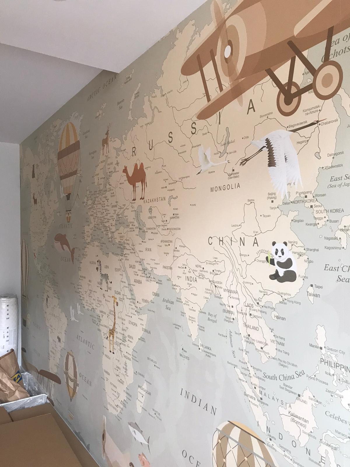 Ściana w pokoju dziecięcym pokryta tapetą z mapą świata, z ilustracjami zwierząt, samolotu i balonu na ogrzane powietrze, oraz naklejonym pandą i żurawiem.