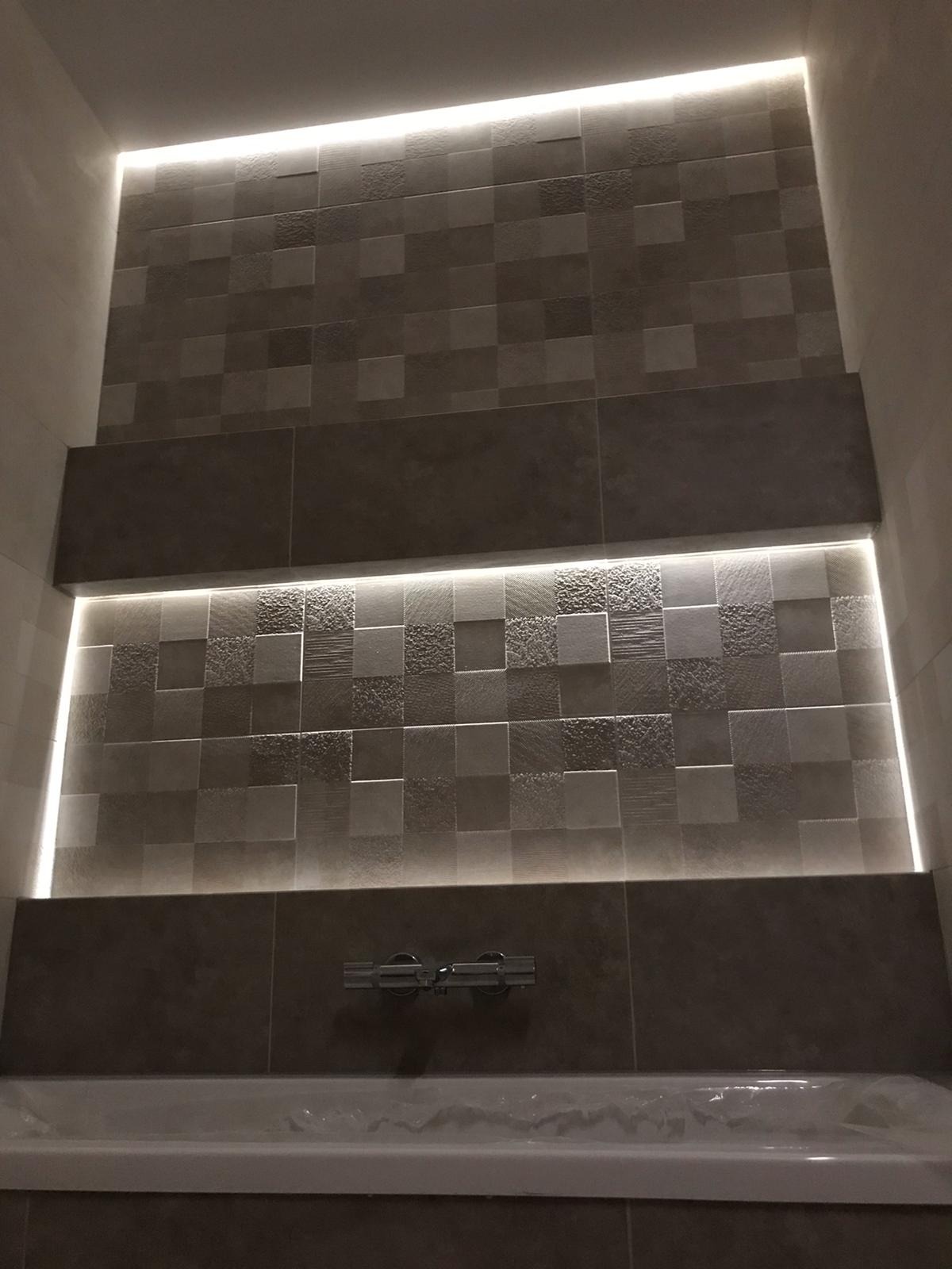 Ściana nad wanną wyłożona mozaiką z szarych płytek o zróżnicowanej fakturze, podświetlona ledowym paskiem, widoczna bateria wannowa.