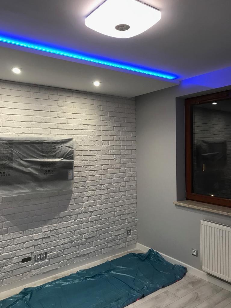 Pokój z białą ceglaną ścianą, zabezpieczoną folią podłogą, oświetleniem LED pod sufitem i oknem z brązową ramą.