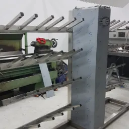 Metalowa konstrukcja z ramionami i czarnymi nakładkami, prawdopodobnie stojak lub wózek warsztatowy, w tle widoczna tokarka konwencjonalna.