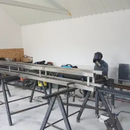 Pracownik w masce spawalniczej łączy metalowe profile na kozłach w warsztacie.