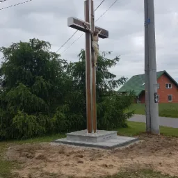 Drewniany krzyż z figurą Chrystusa na kamiennym postumencie, obok drogi i słupa energetycznego, w tle dom z zielonym dachem.