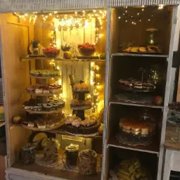 Rustykalna witryna wypełniona różnorodnymi deserami: ciastami, babeczkami, owocami w szklanych naczyniach i słoikach, oświetlona ciepłymi lampkami, tworząca świąteczny klimat bufetu.