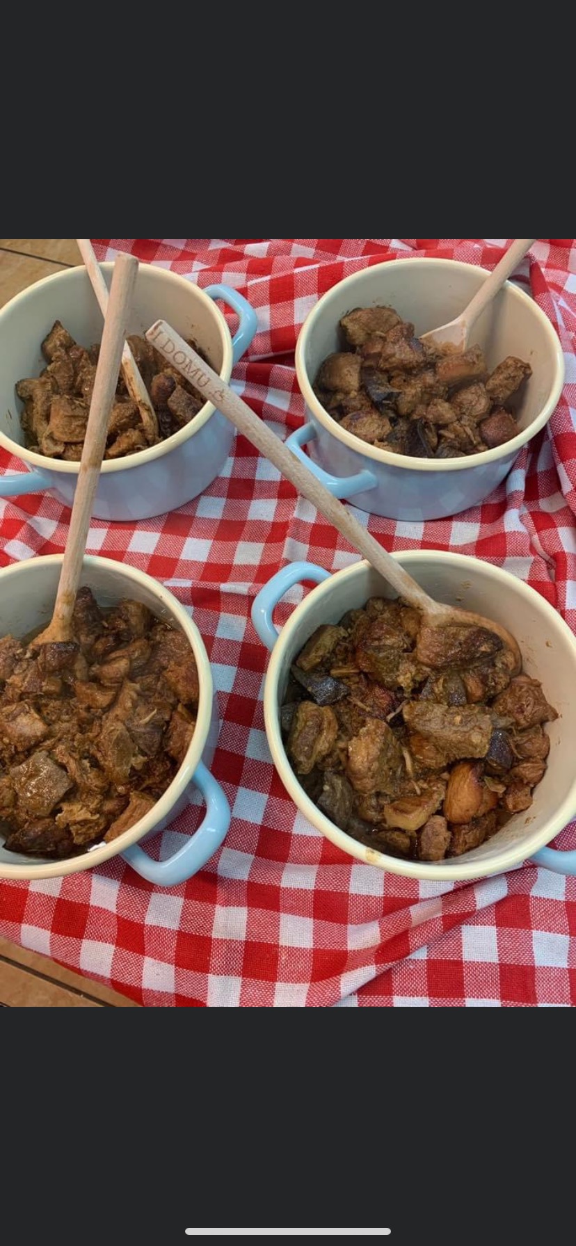 Cztery porcje gulaszu w jasnych miseczkach z drewnianymi łyżkami na czerwono-białym obrusie w kratę. Łyżki mają wygrawerowane logo 'DOMU'.