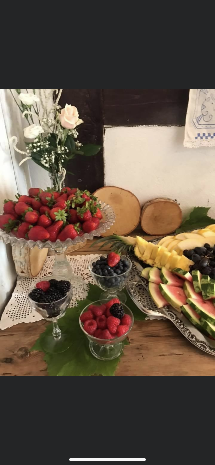 Dekoracyjny stół z owocami: truskawki w szklanej misie na nóżce, maliny, jeżyny i borówki w kieliszkach, plasterki arbuza i ananasa na srebrnej tacy, całość uzupełniona kwiatami i drewnianymi...