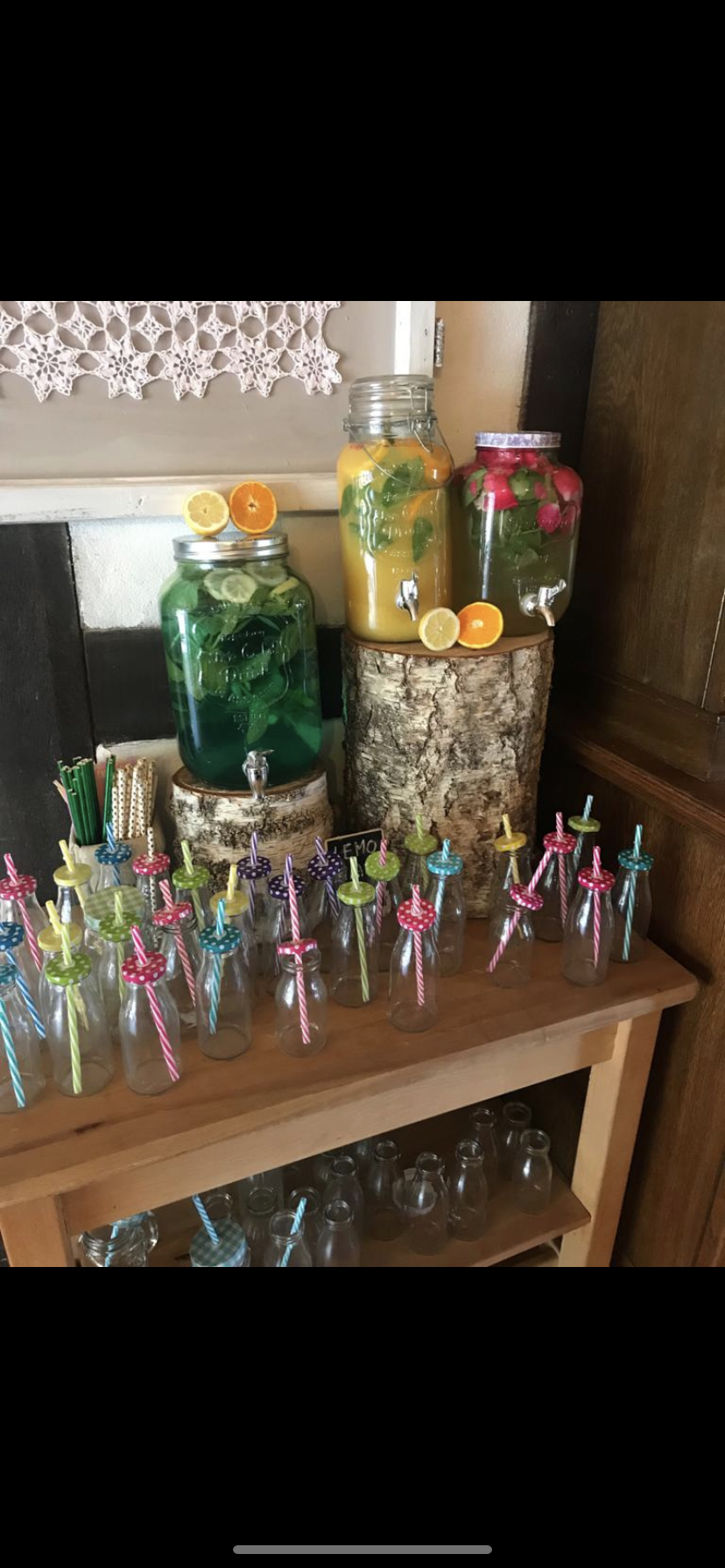 Przygotowany bufet z napojami: trzy szklane słoje z kranikami wypełnione kolorowymi napojami owocowymi i ziołowymi, ustawione na drewnianych pniakach, a przed nimi rząd szklanych butelek...