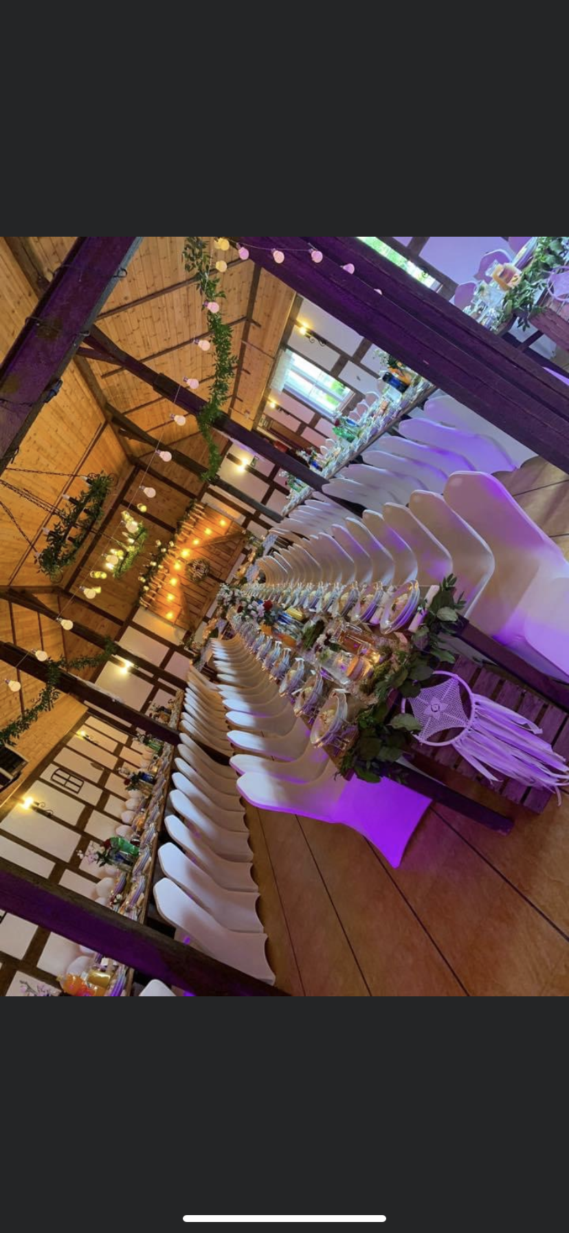 Ustawiony długi stół z białymi krzesłami i dekoracjami kwiatowymi w rustykalnej sali z drewnianą konstrukcją dachu i girlandami świetlnymi.