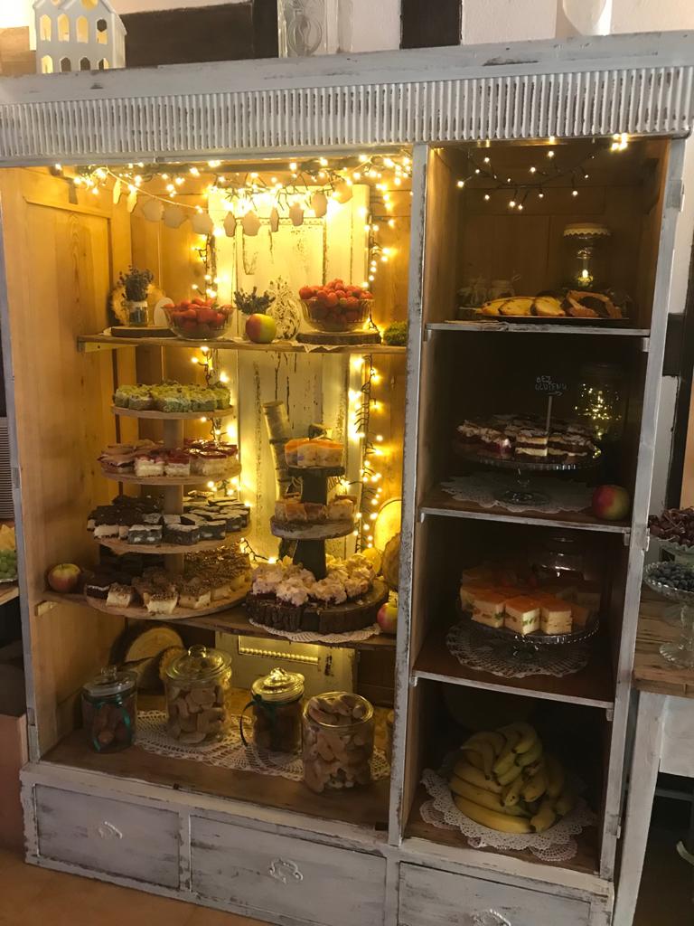 Rustykalna witryna wypełniona różnorodnymi deserami: ciastami, babeczkami, owocami w szklanych naczyniach i słoikach, oświetlona ciepłymi lampkami, tworząca świąteczny klimat bufetu.