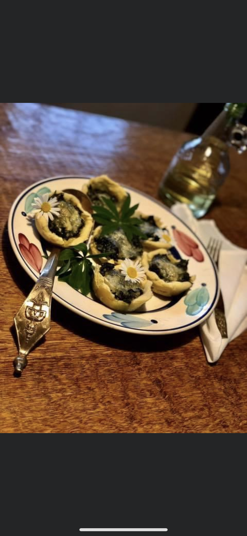 Talerz z mini tartami ze szpinakiem i serem, udekorowanymi świeżymi margaretkami, na drewnianym stole obok butelki z napojem i serwetki, z ozdobnym nożem z motywem roślinnym po lewej stronie.