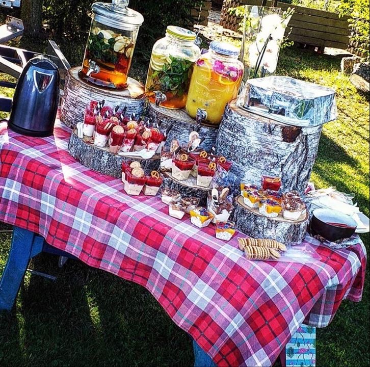 Stół przykryty czerwoną kratą, na nim szklane pojemniki z napojami owocowymi na drewnianych podstawkach oraz desery w plastikowych kubeczkach, część z preclami, na zewnątrz.