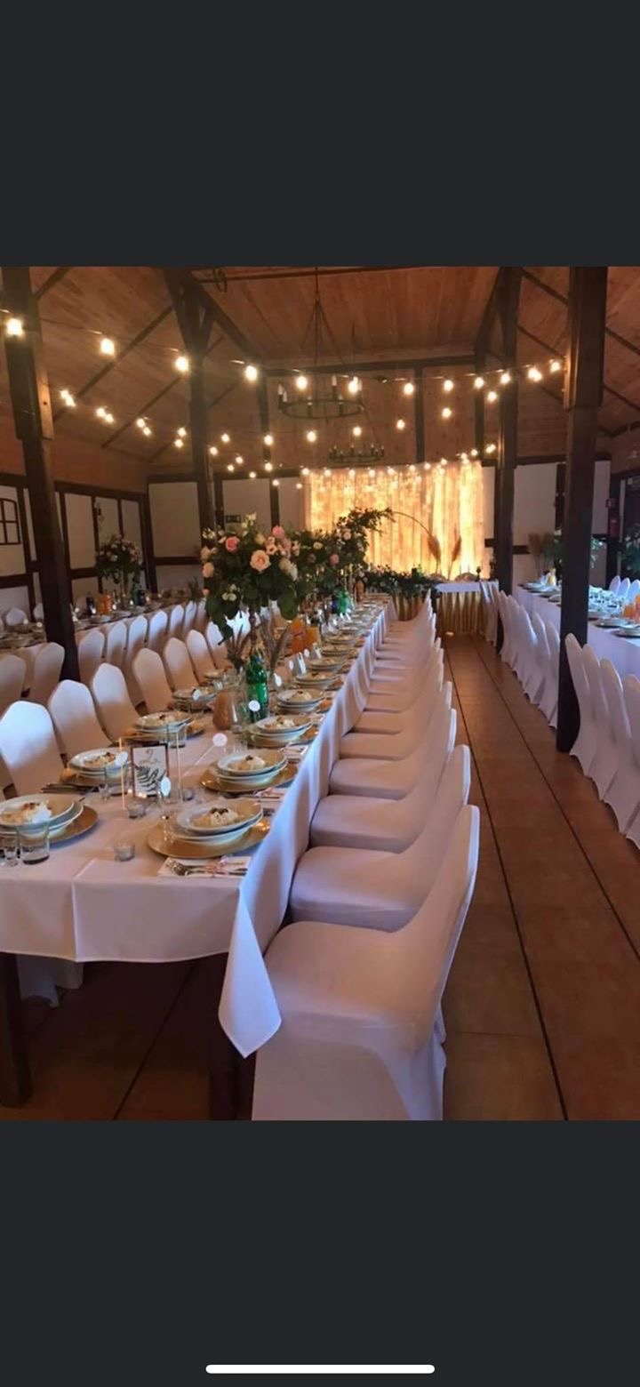 Uroczysta sala bankietowa z długim stołem nakrytym białym obrusem, krzesła w pokrowcach, dekoracje kwiatowe i świetlne, rustykalny wystrój wnętrza z drewnianym stropem.