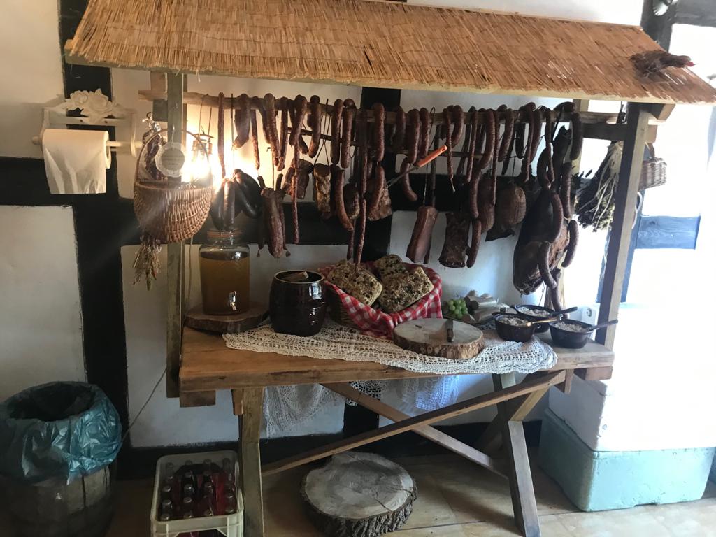 Stoisko z wędlinami i pieczywem pod strzechą, rustykalny wystrój, słoik z kranikiem, gliniane naczynia, dekoracyjna serweta, koszyk z pieczywem w czerwoną kratę.