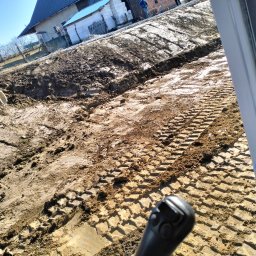 MAJKI-KOP - Świeżo wykopany rów w ziemi z widocznymi śladami opon maszyny budowlanej i fragmentem białej rury, widziany z perspektywy operatora maszyny.