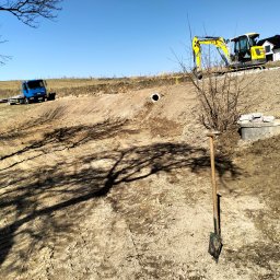 MAJKI-KOP - Żółta koparka na zboczu z widocznym wykopem, rurą drenażową i studzienką betonową, w tle niebieska ciężarówka i dom, cień drzewa na pierwszym planie, łopata wbita w ziemię.