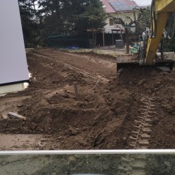 MAJKI-KOP - Żółta koparka w trakcie prac ziemnych, widoczne świeże ślady gąsienic na wyrównanym terenie, w tle dom z panelami słonecznymi i wysokie drzewa iglaste. Widok z kabiny operatora.