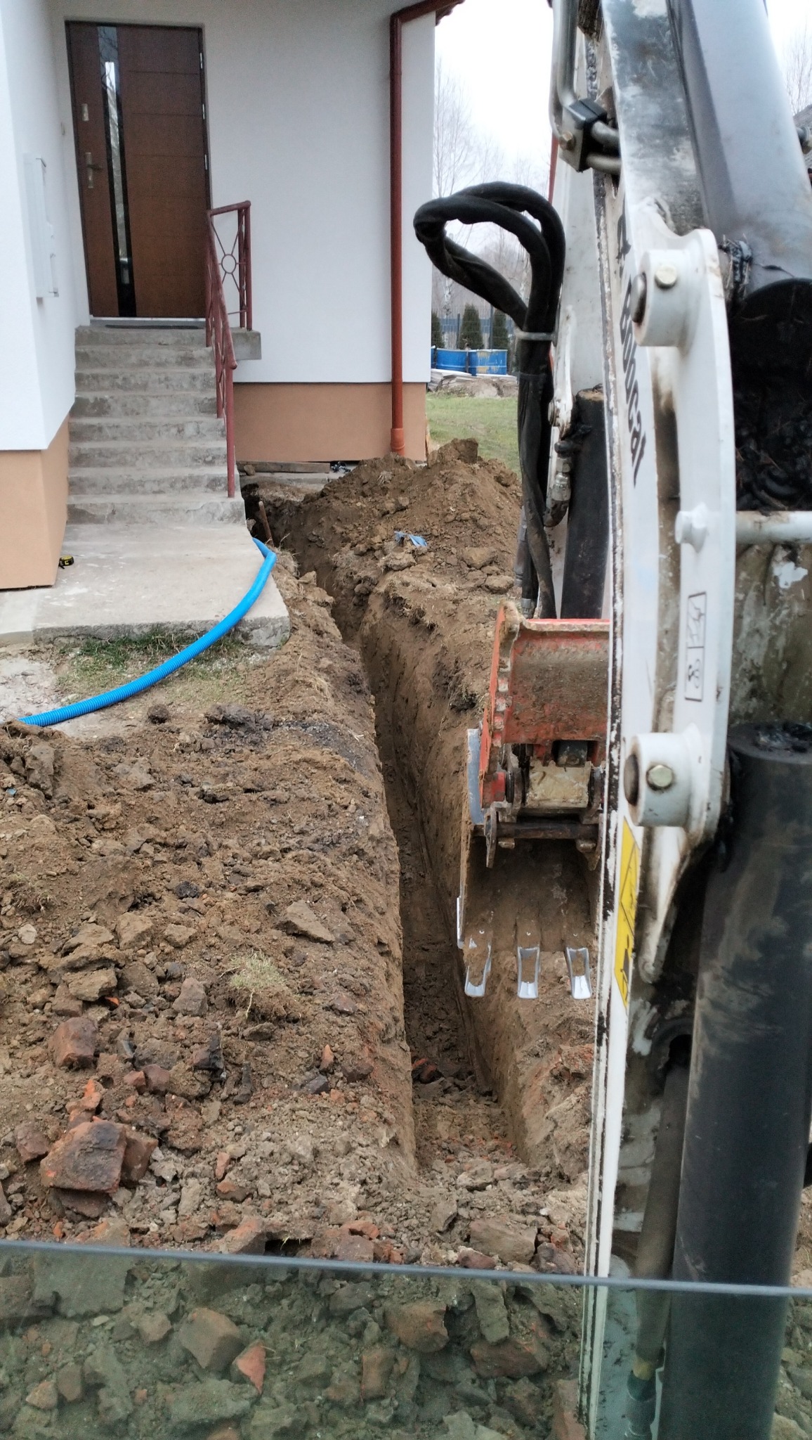 Wąski wykop liniowy przy fundamencie domu, wykonany koparką Bobcat, widoczny niebieski przewód w pobliżu