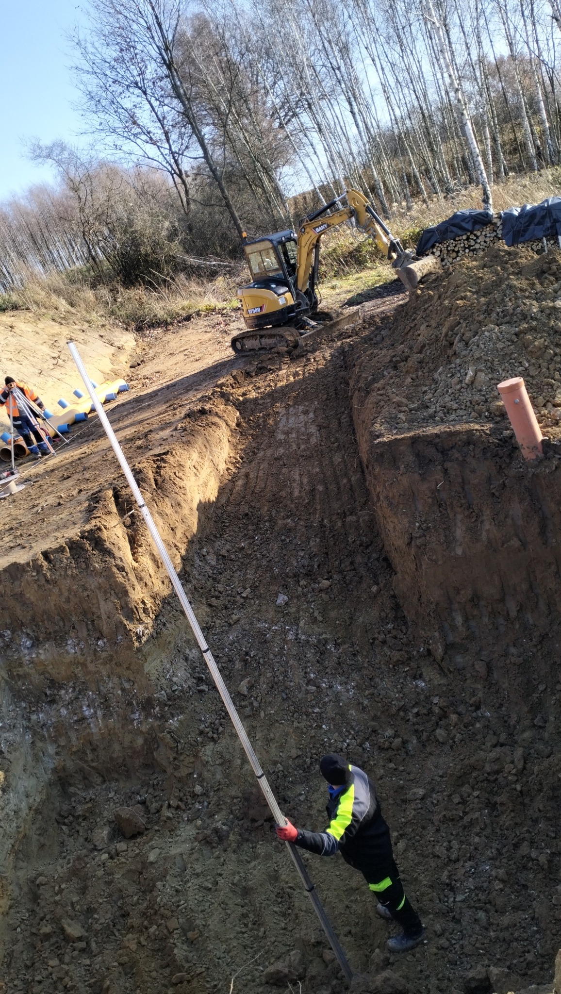 Żółta minikoparka SANY na skraju wykopu, w tle ułożone drewno pod plandeką, pracownik z łatą niwelacyjną mierzy głębokość wykopu, w oddali widoczny niwelator i rury.