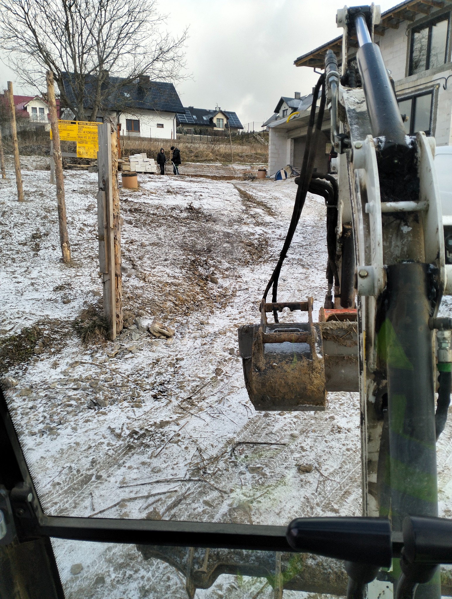 Widok z kabiny minikoparki na częściowo odśnieżony teren budowy z drewnianymi słupkami, w tle domy i dwóch pracowników.